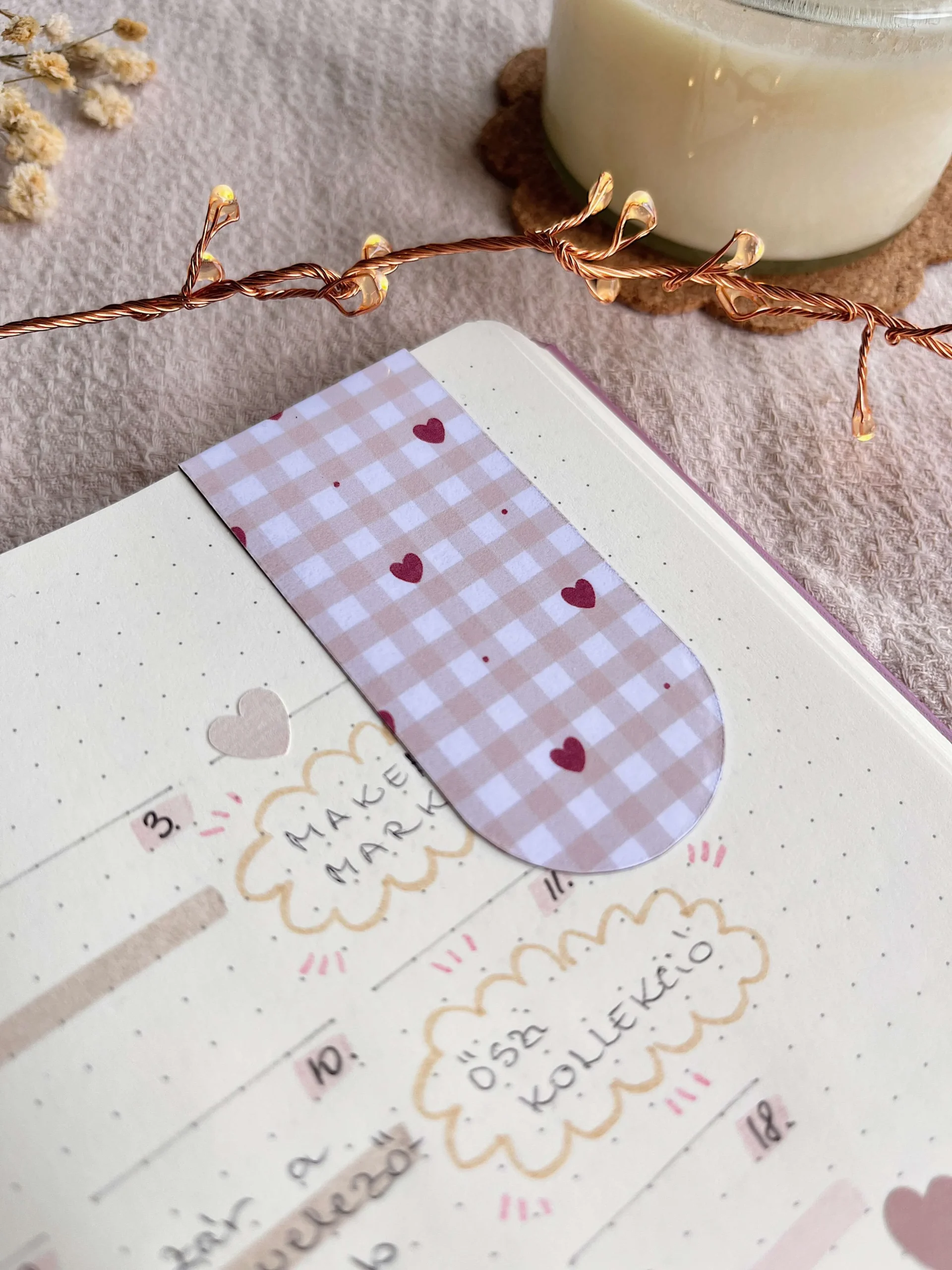Barna gingham és szívecske mintás mágneses könyvjelző naplóban, körülötte hangulatos fények, gyertya és virág. Cozy olvasós élmény.